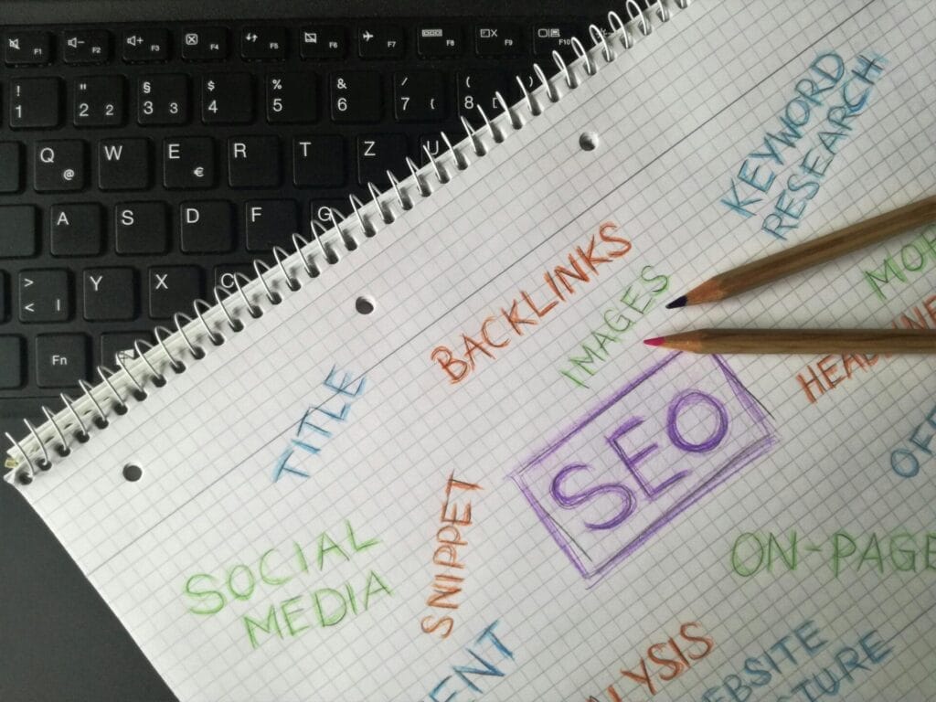 Auf einer Laptop-Tastatur liegt ein Notizblock mit mit Buntstift geschriebenen SEO-Schlüsselwörtern, auf der Seite liegen zwei Bleistifte.