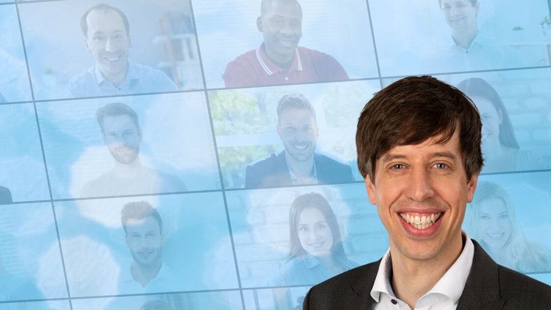Im Vordergrund lächelt ein Mann im Anzug, hinter ihm sind auf einem unscharfen virtuellen Meeting-Bildschirm viele Menschen zu sehen.