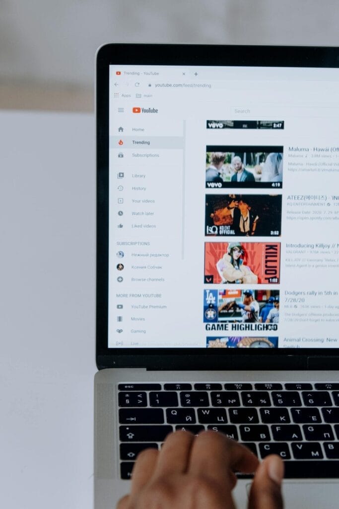 Die Hand einer Person, die auf einem Laptop tippt, während die YouTube-Homepage geöffnet ist, um YouTube-Videos zu durchsuchen und verschiedene Miniaturansichten und Navigationsoptionen zu erkunden.